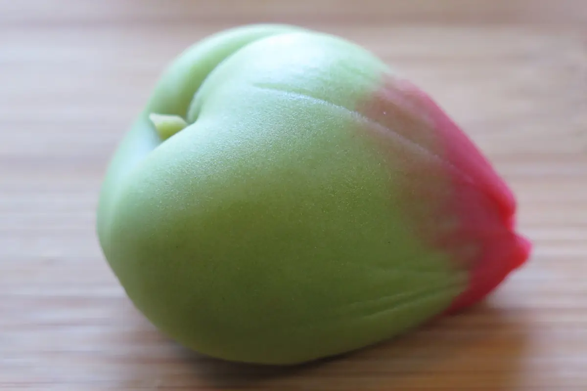 A pale yellow wagashi sweet shaped like a flower with a brown centre and a green leaf, on a wooden table