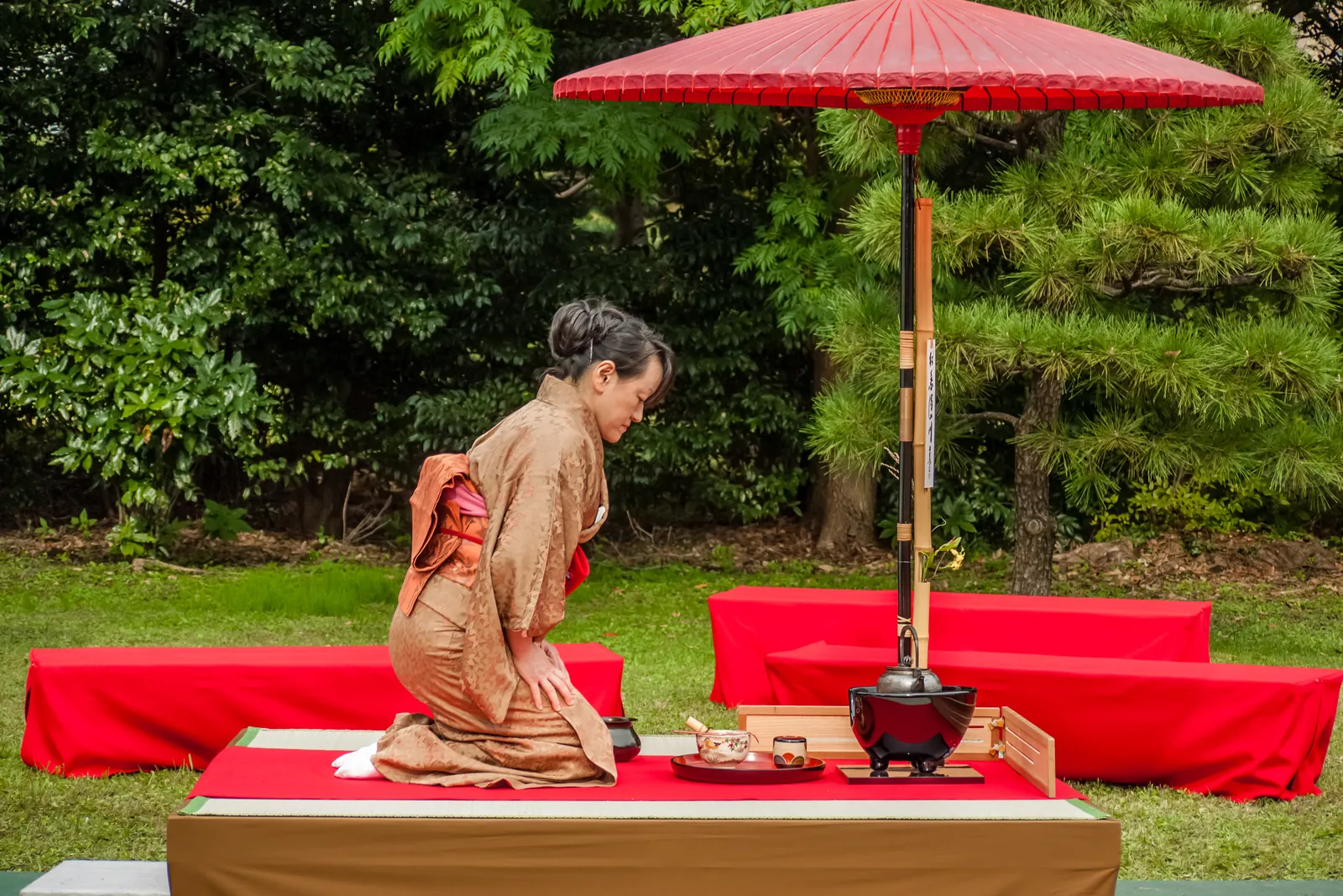 A woman in a brown kimono kneels at a tea station under a red parasol in a garden, an iron kettle and tea utensils in front of her; a tea host at an outdoor gathering in Tokyo, not at this tea house
