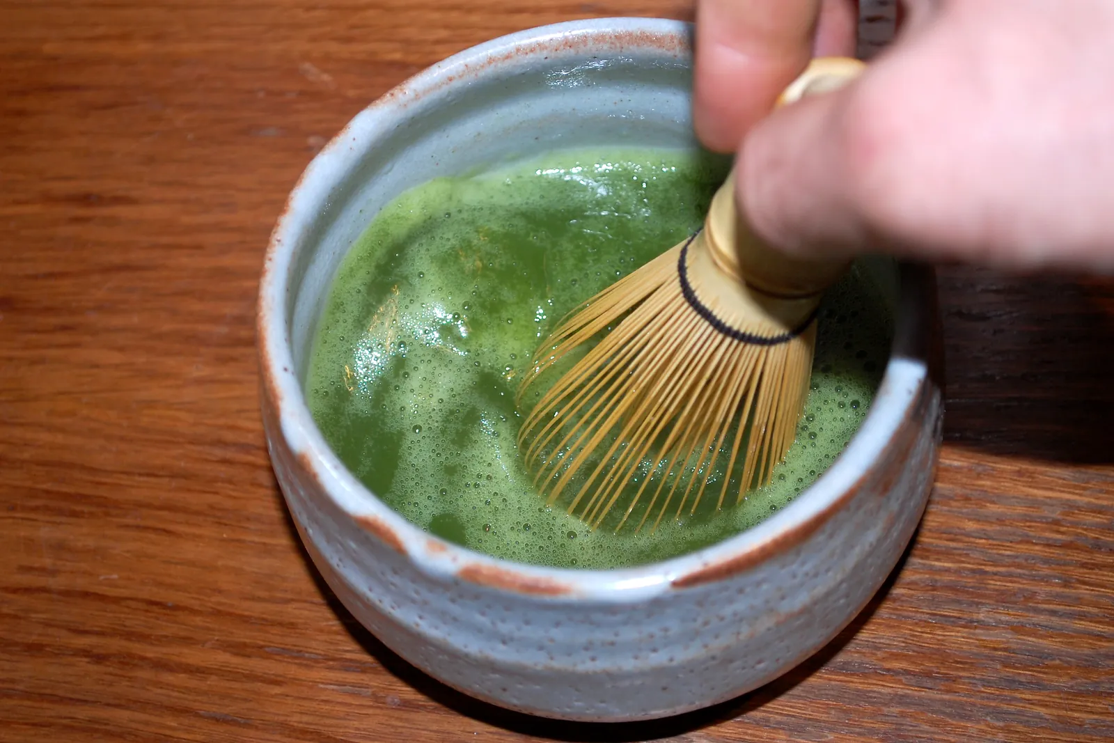 A hand turns a bamboo whisk in a bowl of frothy green matcha; a general photograph of preparing matcha, not of this tea house